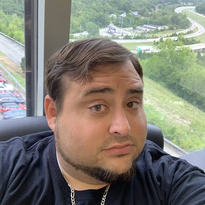 Man in dark shirt with silver chain necklace in high-rise office with expansive view of Cincinnati valley, parking lot, and green landscape below