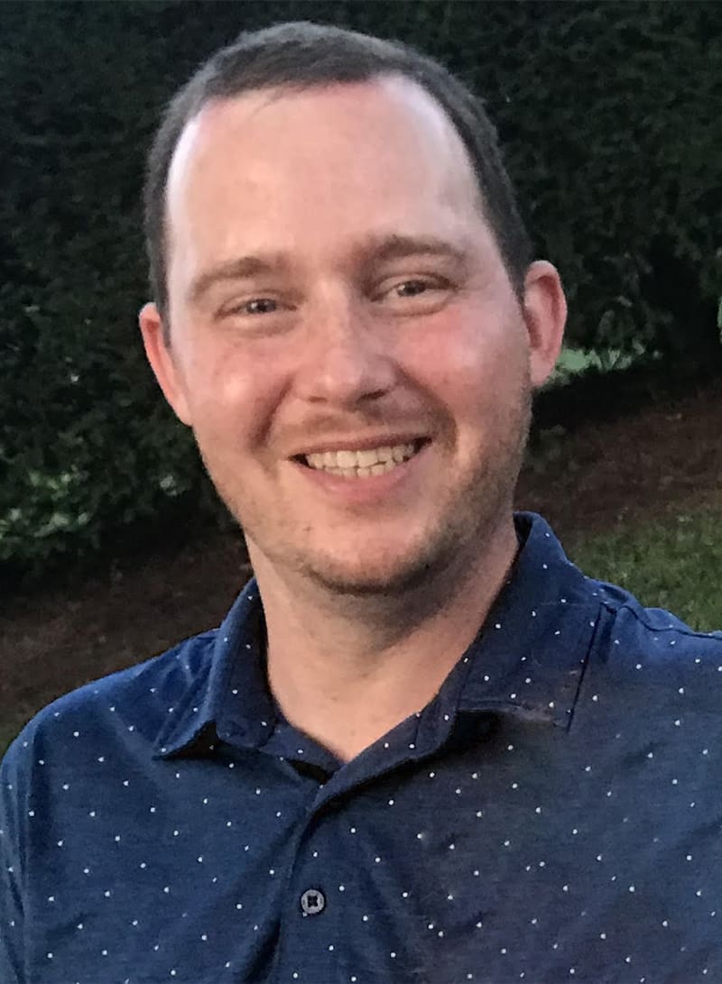 Smiling man wearing a dark blue button-up shirt with white speckles, photographed outdoors against a green hedge background