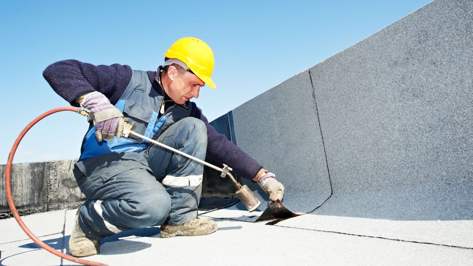 Roofing contractor wearing a yellow hard hat and protective gear applies a torch-down membrane to a flat commercial roof for waterproofing. Empire Contractors specializes in professional flat roof installation and repair for long-lasting durability and weather resistance.