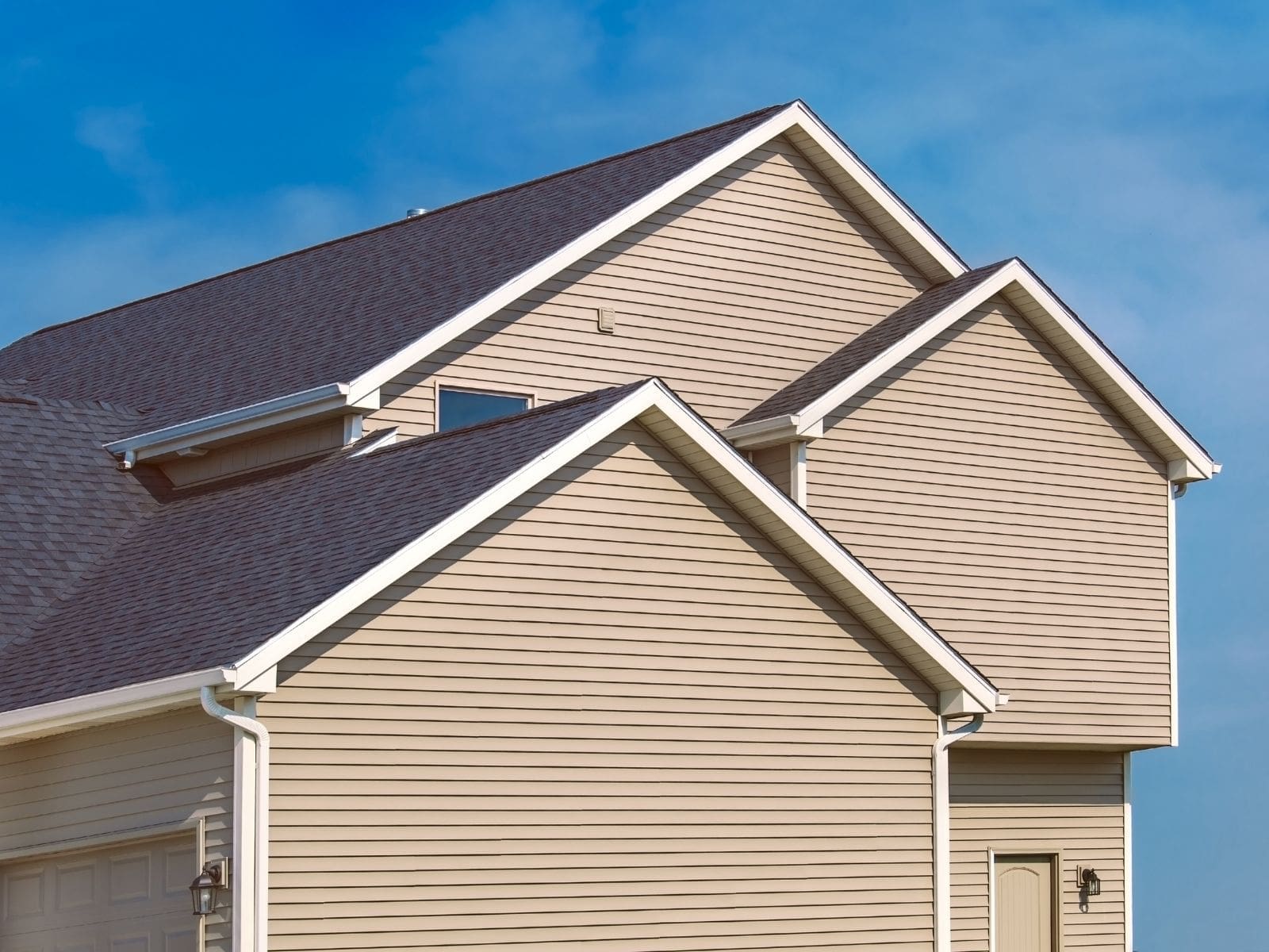 Modern home with beige vinyl siding, dark asphalt shingle roof, multiple gables, and attached garage under clear blue sky