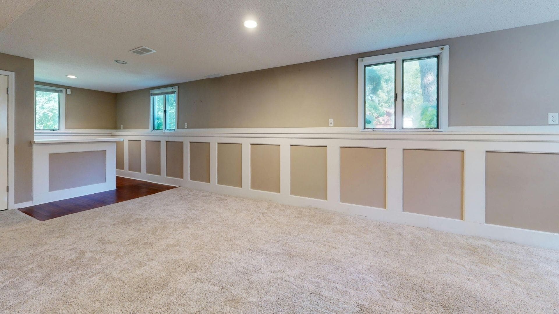Spacious basement room with beige walls, white wainscoting panels, carpet flooring, large windows, recessed lighting, and a fireplace accent wall